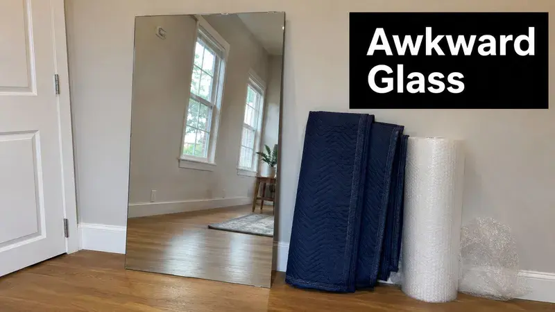 A large rectangular mirror leans against a wall next to moving blankets and a roll of bubble wrap.