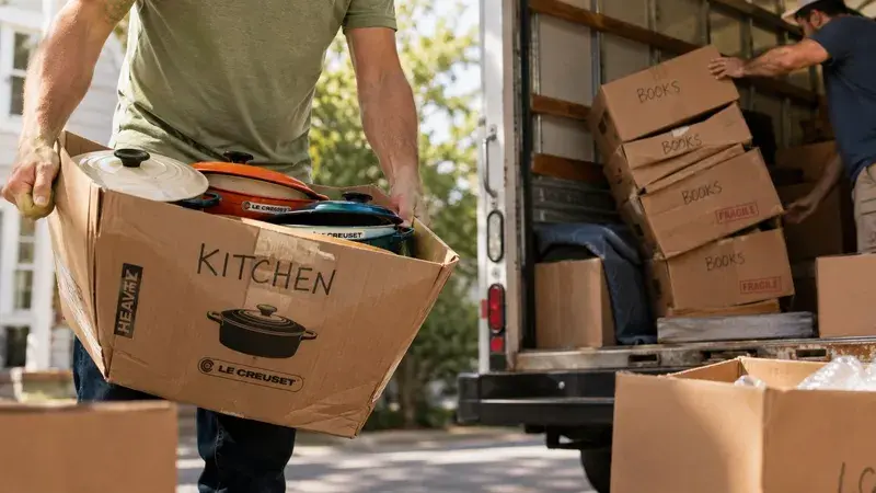 A person carrying a heavy box labeled kitchen filled with Le Creuset cookware during a residential move.